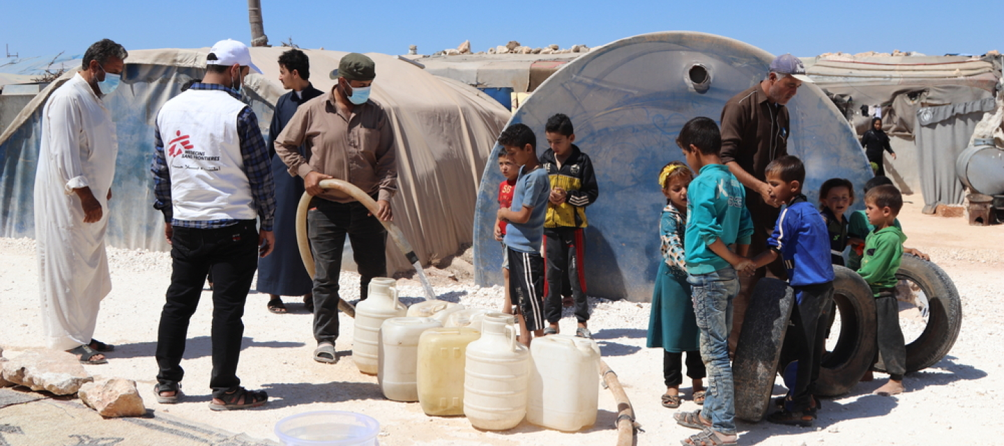 Mitarbeiter von Ärzte ohne Grenzen befüllen Wasserkanister, daneben eine Gruppe von Kindern
