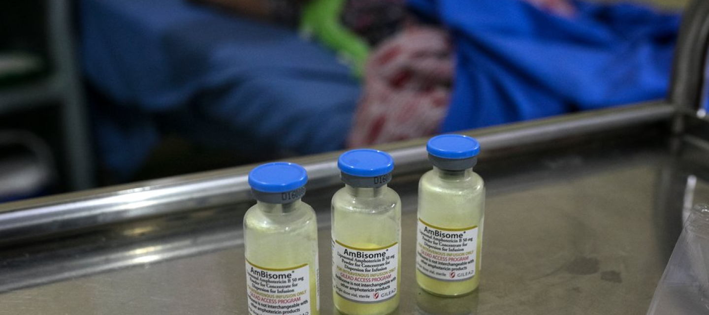 Drei Impfampullen stehen auf einem Tisch in einem Krankenhaus. Im Hintergrund ist eine Patientin zu sehen.