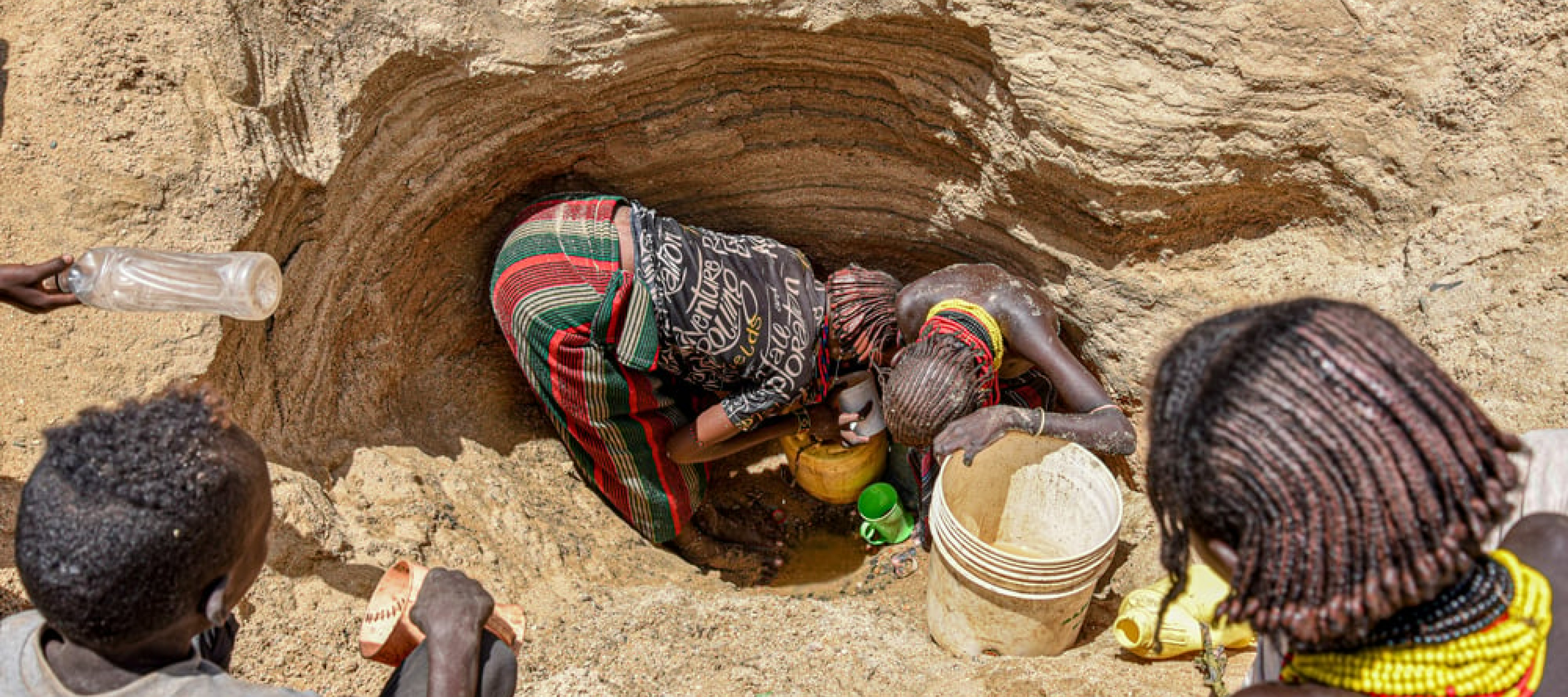 Zwei Frauen stehen in einem Loch im Boden und schöpfen Wasser, andere außenherum sehen zu